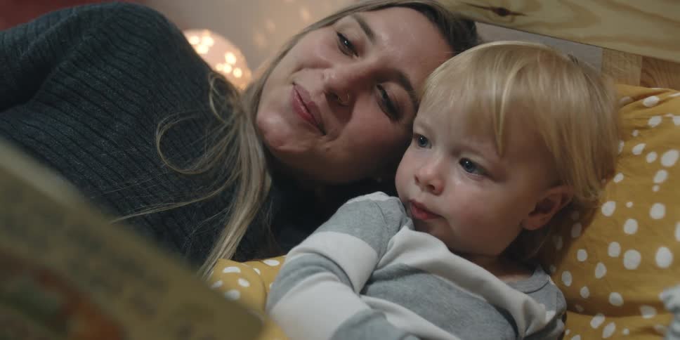 A woman reads a book to a toddler while lying beside him in bed