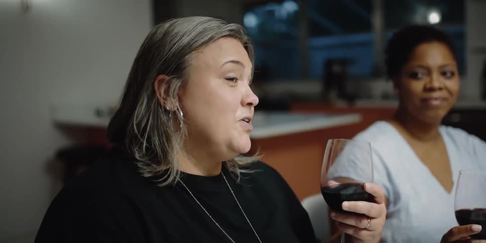Two women hold goblets of wine while one of them talks and the other watches