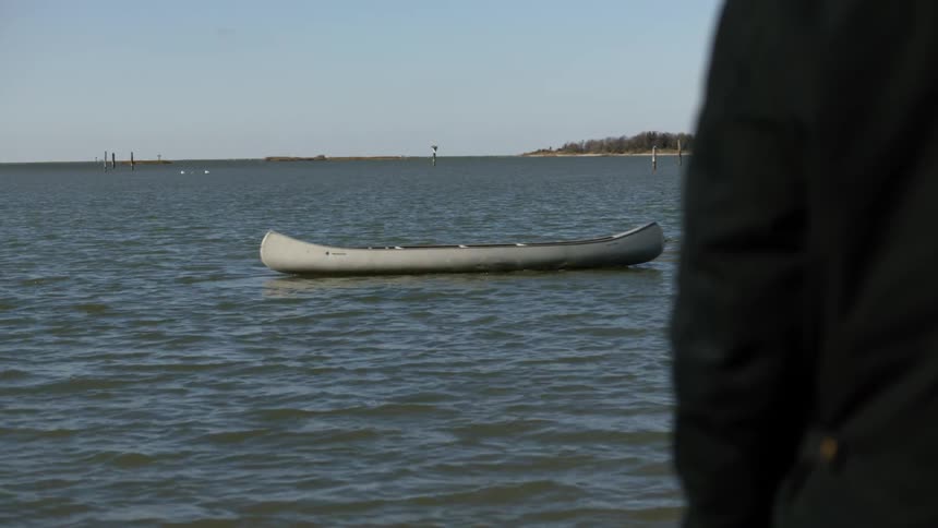 An empty canoe is moved by something underwater as someone watches