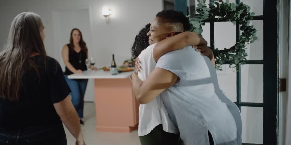 A woman walks in to a home and hugs another woman as others walk to greet her