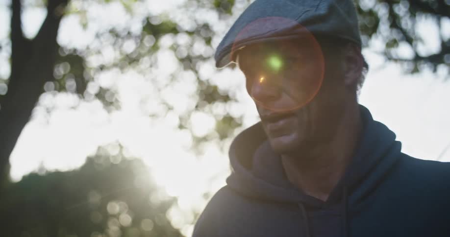 A man wearing a hat and hoodie stands amidst trees on a sunny day