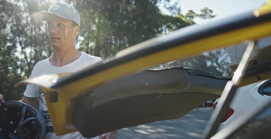 A man with a hat closing the back of the vehicle while holding a helmet and inline skates with trees the sky and clouds in the background