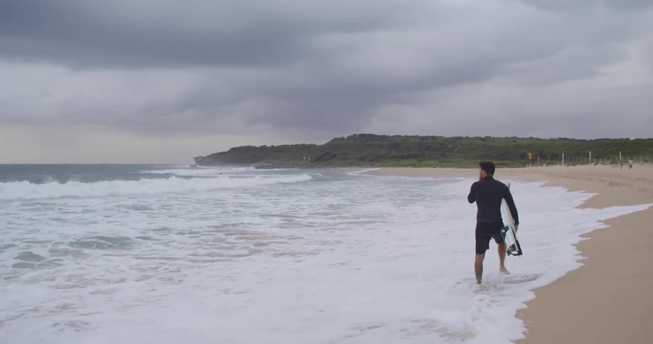 Man wearing a black wetsuit and carrying a surfboard is walking across the beach watching the waves