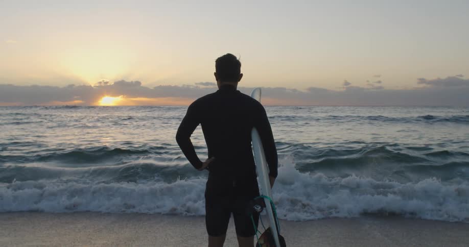 Surfer stands on beach holding surfboard watching sunrise on horizon