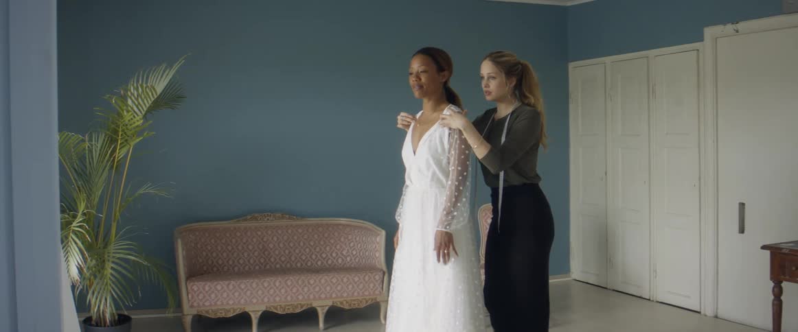 Two women in a room with one with a measuring tape and adjusting a dress on the other woman with a wall in the background