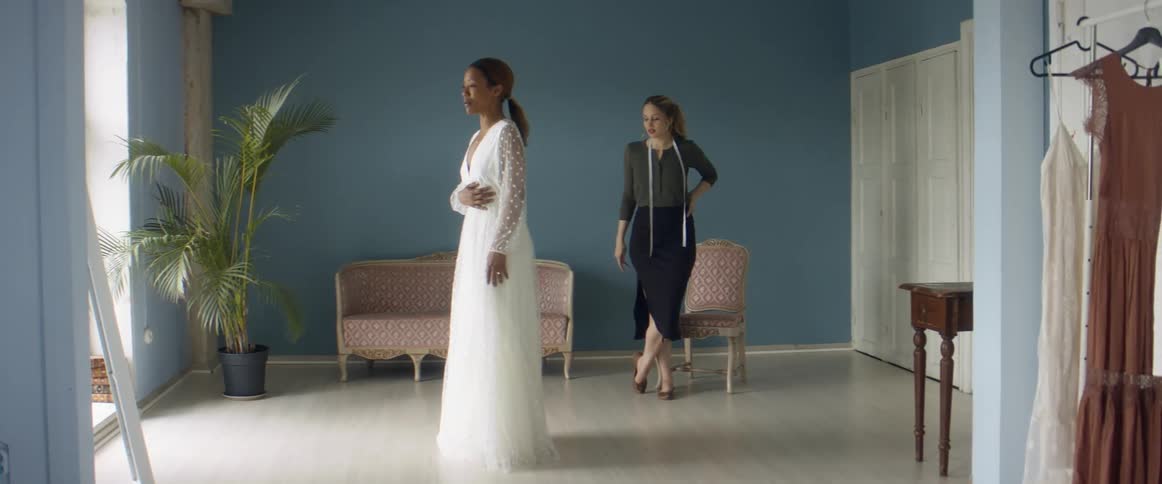 Two women in a room with one looking at a dress in a mirror with a wall and window in the background
