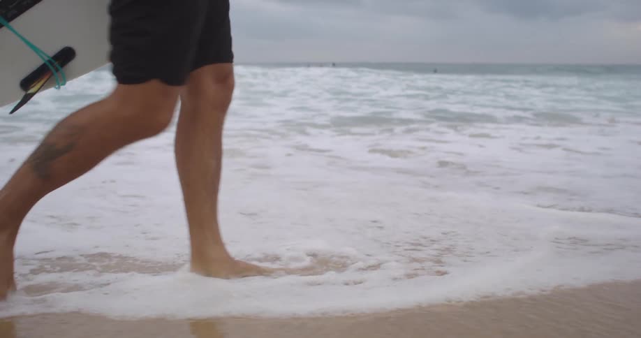 Barefoot man wearing shorts and carrying a surfboard is walking across a beach as water rolls in over his feet