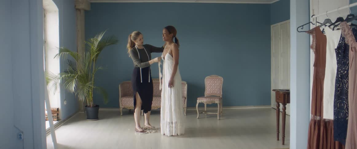 A tailor holds up lace trim to a dress being worn by a woman studying herself in a mirror