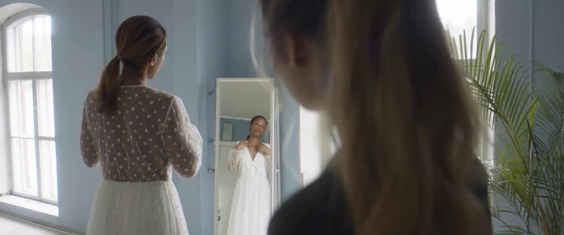 Two women in a room with one looking at a dress in a mirror with a wall and window in the background