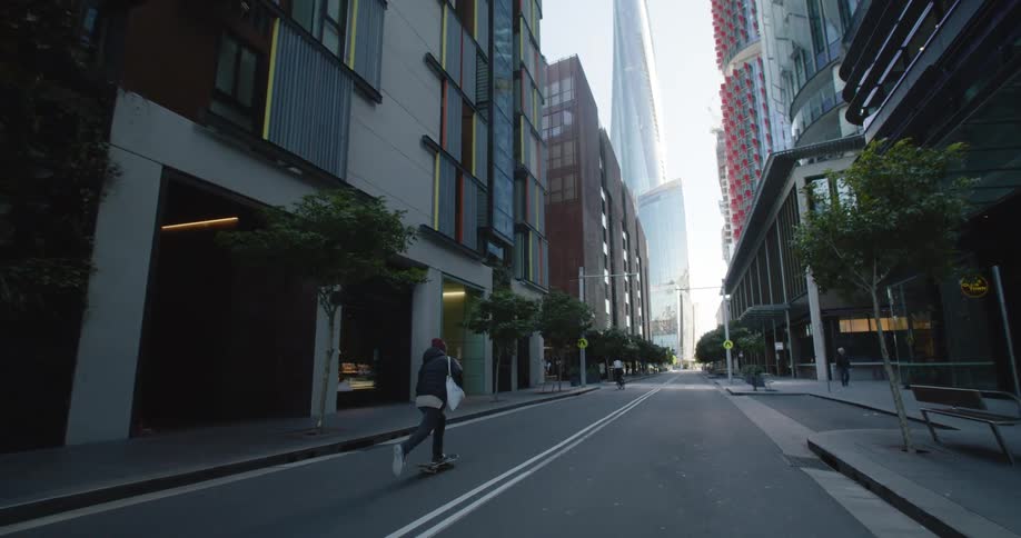 Man with beanie black coat and white bag skateboards in the city street with high rise buildings