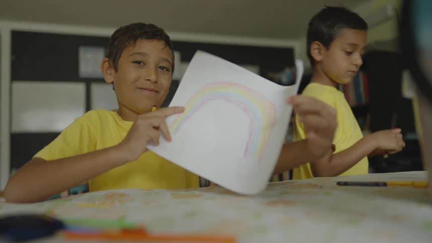 Two boys in a classroom with one holding a paper and pencil with a wall in the background