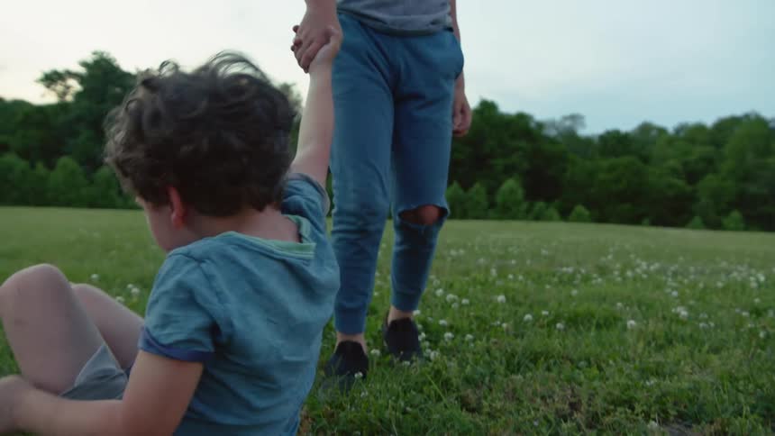 A little boy laying down in a grass field is helped up off the ground by his brother during the day