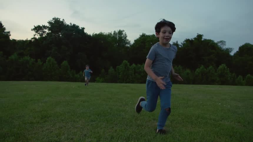 Two boys run in the grassy field by the forest