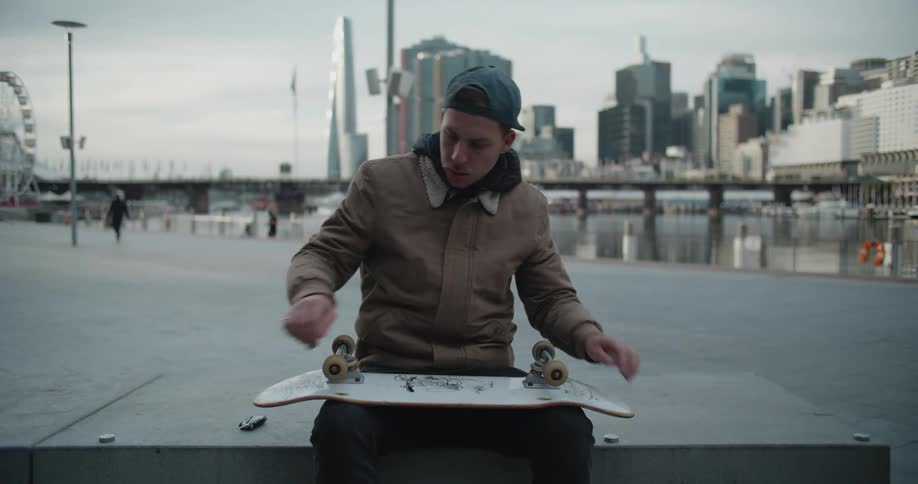 A man sitting on a bench holding a skateboard with buildings the sky and clouds in the background