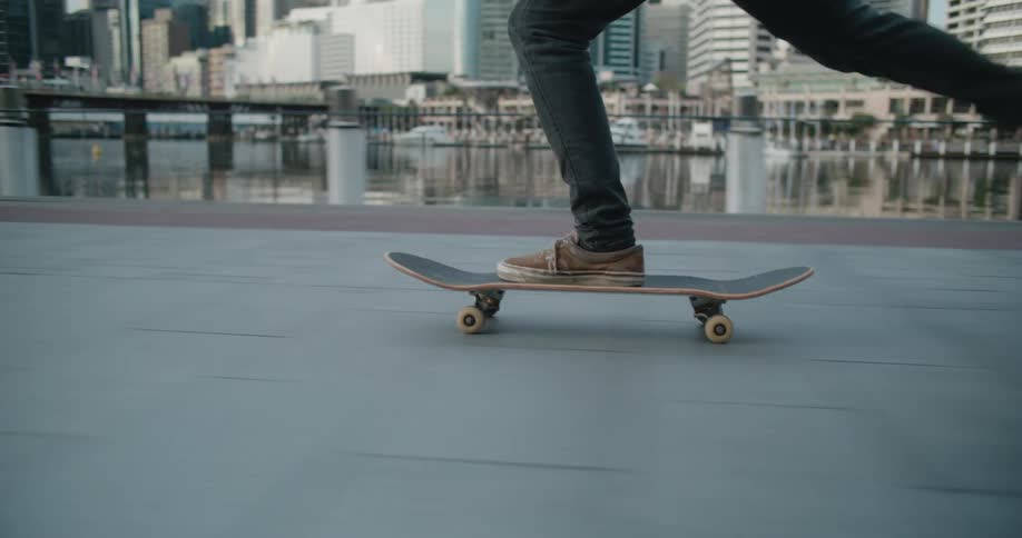 Man with brown shoes and jeans skateboards next to river with high rise buildings in the background