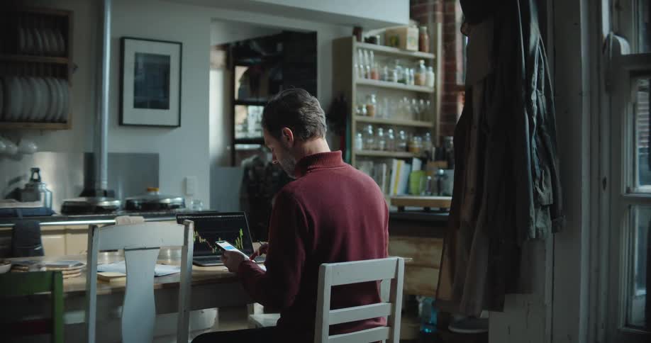 A man is working in his home using a laptop and smartphone while sitting at his kitchen table