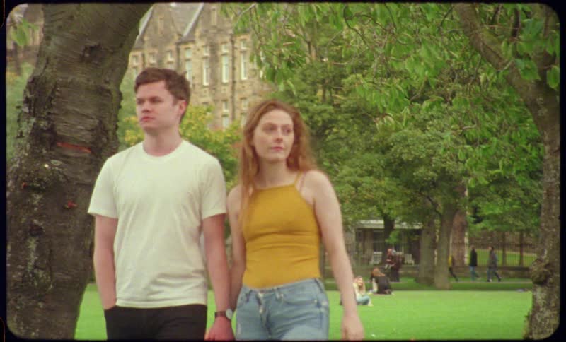 Couple wearing short sleeves walks forward as people wearing cold weather gear walk backward near wide lawn bordering buildings