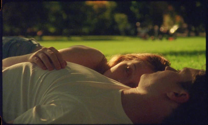 A man and the woman laying in grass with trees and vehicles in the background