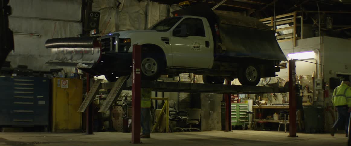 Mechanic works on truck with attached snow plow sitting on lift in car garage