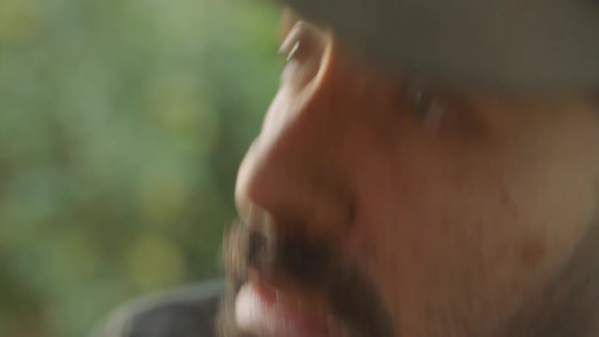 A man is standing outdoors by plants and wearing a hat