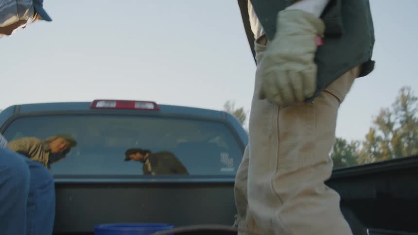 Man carries bucket to truck parked near trees before climbing into pickup truck bed and sitting