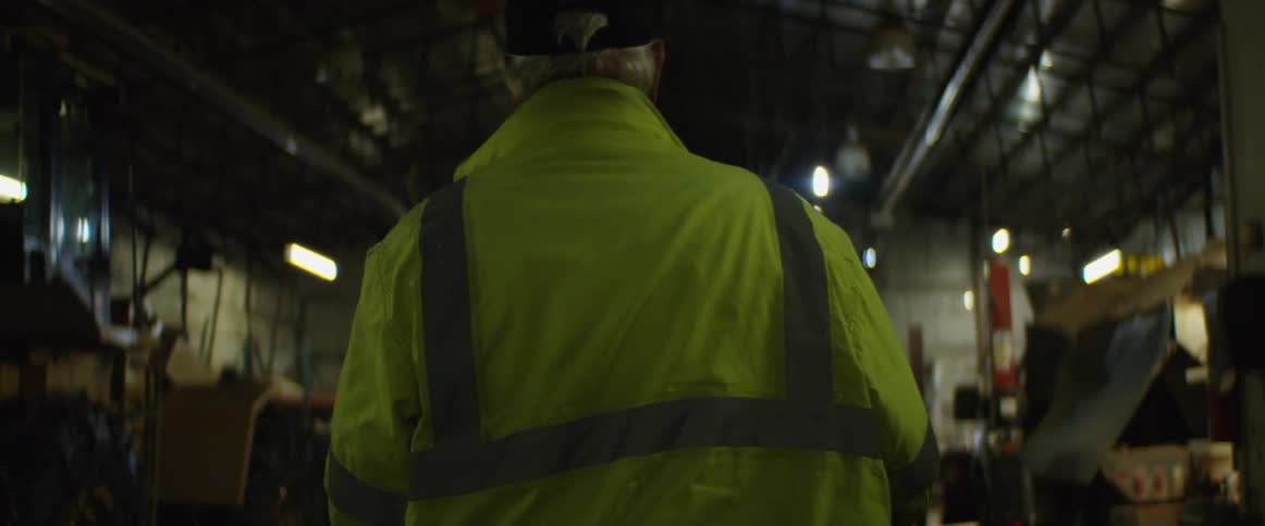 Older man wearing safety jacket walks through large storage building toward large vehicle