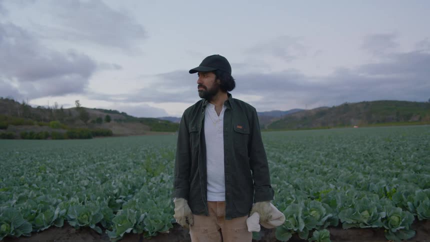 A man with a hat gloves and a bag standing with a field of crops the sky and clouds in the background
