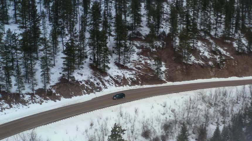 A single black suv drives on a cliffside road way next to mountains covered in snow during the day