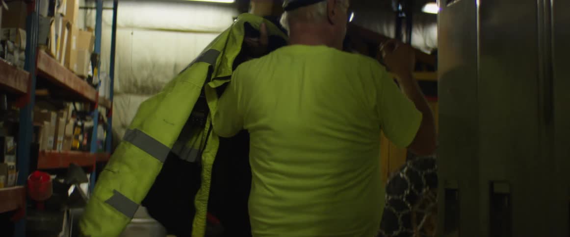 A man opens a locker and takes out a safety jacket that he puts on while walking inside of a warehouse
