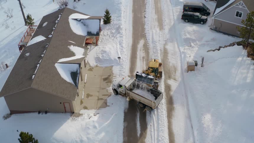 A bulldozer putting snow in the back of a truck in a neighborhood with houses and trees