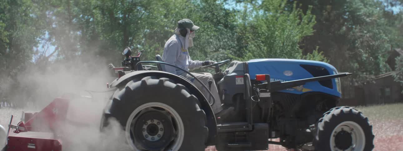Man wearing a cap grey sweatshirt and pants drives blue tractor plowing the field