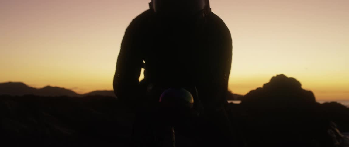 A person with a flight suit and a helmet on a beach with the sky and clouds in the background