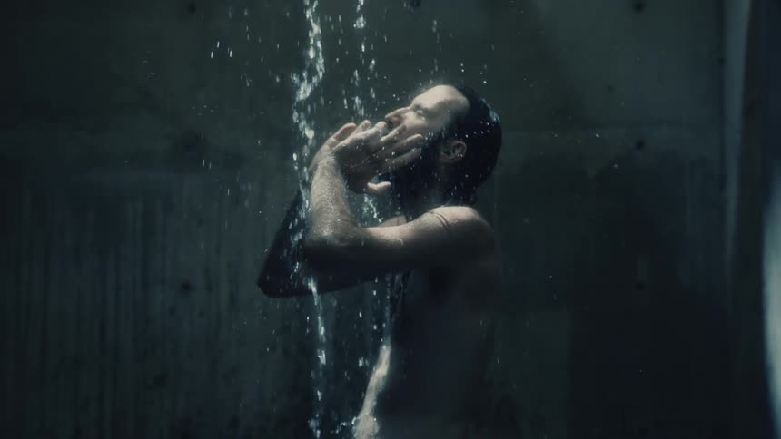 A man stands under a shower of water in a room with concrete slab walls