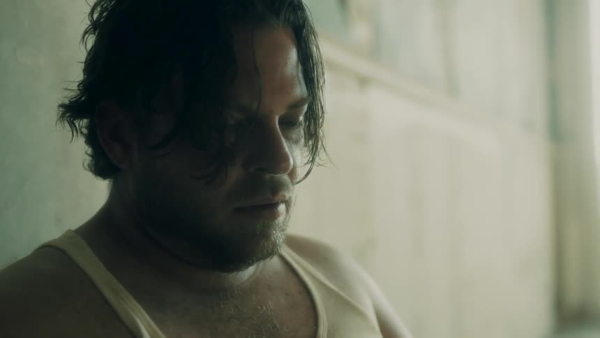 An overweight man in a tank top sits against a wall and looks dejected