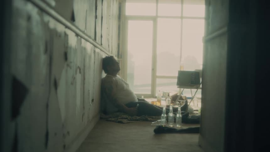 A man sits in a shabby building surrounded by empty beer bottles as he drinks a beer