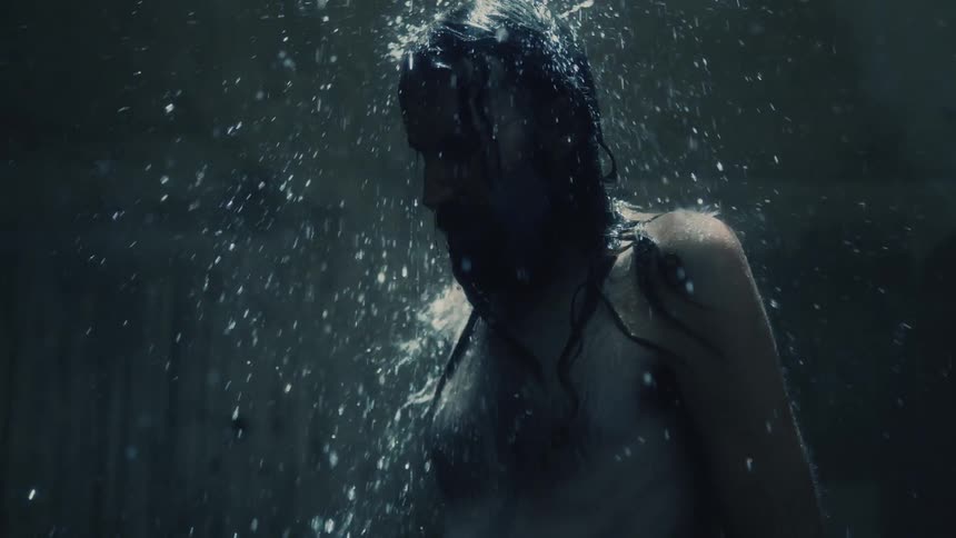 Man shakes his head standing under falling water in dimly lit area with concrete walls