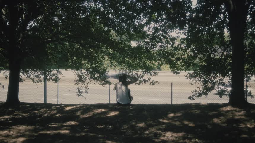 Man sits in shadows of trees overlooking traffic on highway
