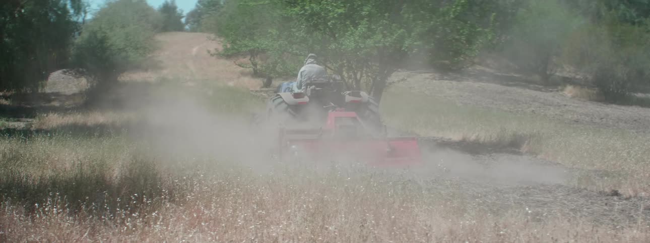 Person wearing over ear headphones drives tractor pulling mowing attachment over grass between trees kicking up dust