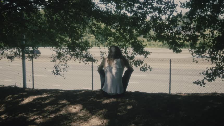 Man sits on ground under trees overlooking traffic on highway