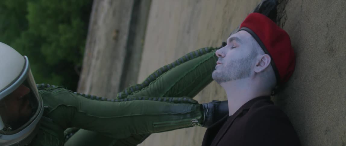 A man wearing a helmet and a green bodysuit eases a dead man in a red beret down on the ground