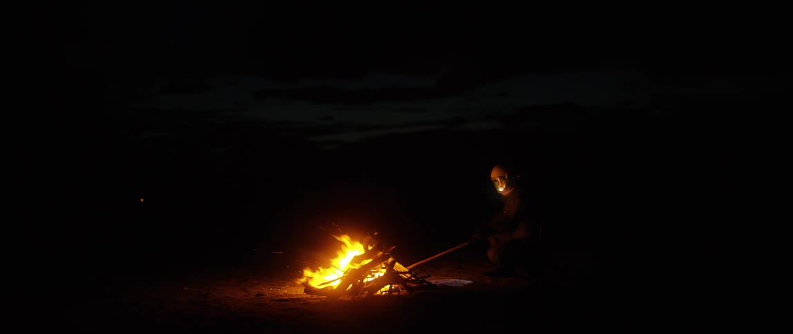 A figure in a body suit and helmet with visor pokes at a camp fire with a stick at night
