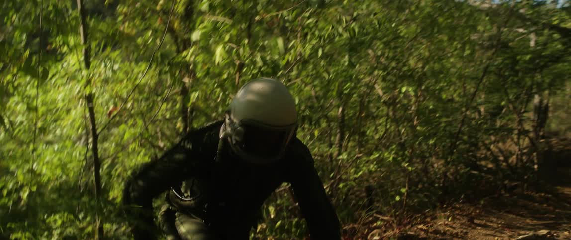 A person with a helmet and flight suit climbing up the hill surrounded with plants and trees with the sky and clouds in the background