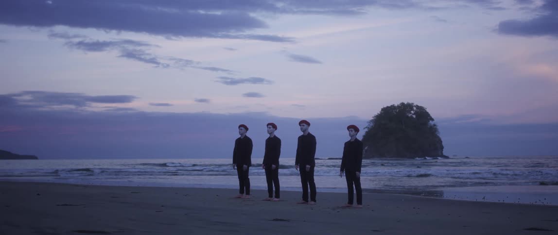 Four men wearing suits and red berets stand barefoot in the sand with the ocean behind them
