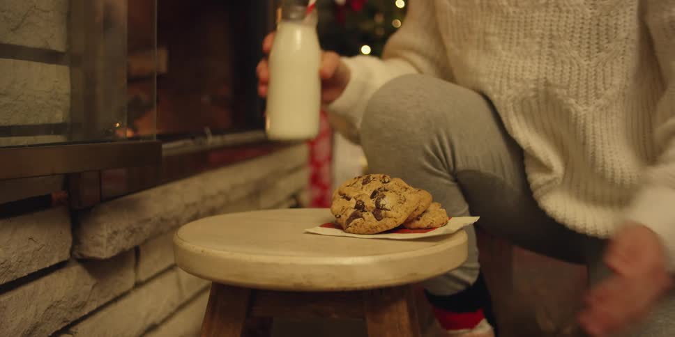 A young woman places milk and cookies on a stool in front of a christmas tree