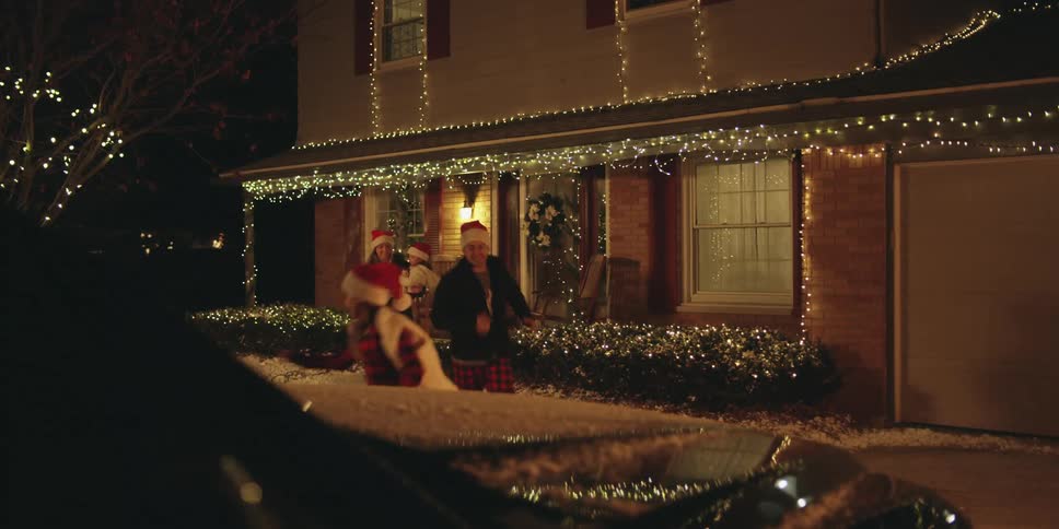 A family leaves a house in matching plaid pajamas wearing santa hats and get in the car