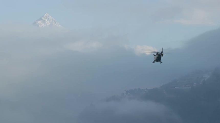 Helicopter flies over fog obscured conifer forest and mountains as mountain summit shows over clouds