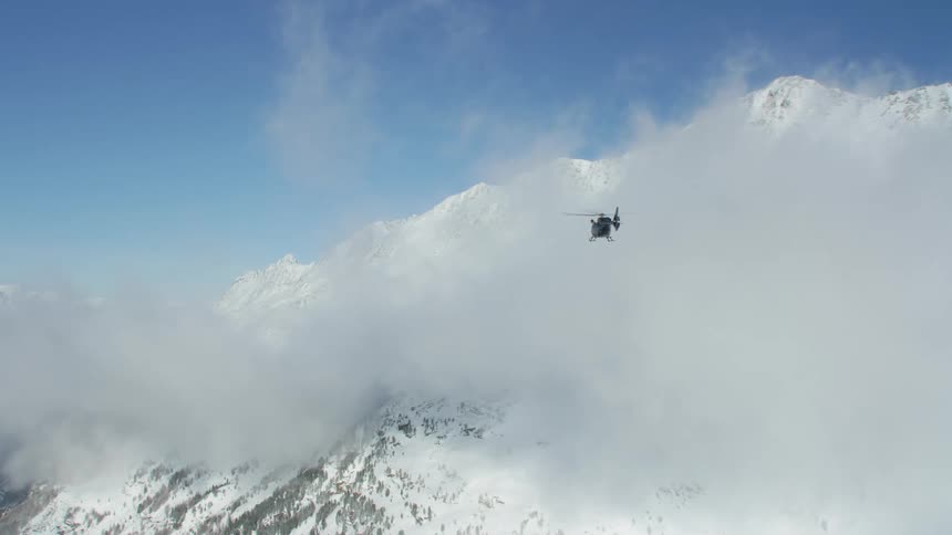 Helicopter hovers near clouds moving over snow and forest covered mountain slope under azure blue sky
