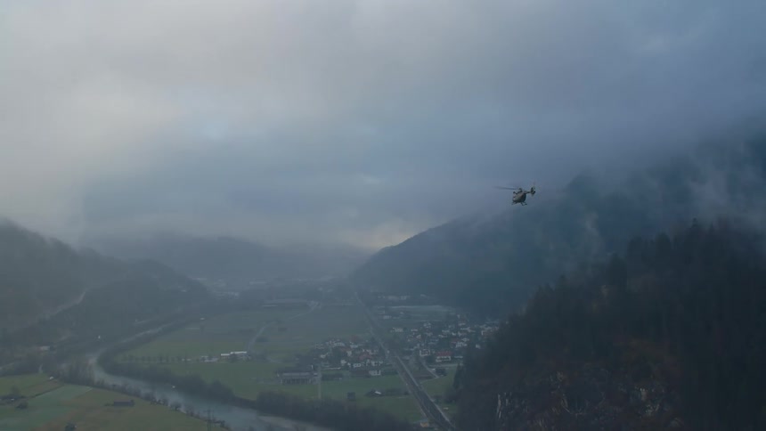 A helicopter flies above a village in a valley near hills and river with thick cloud cover