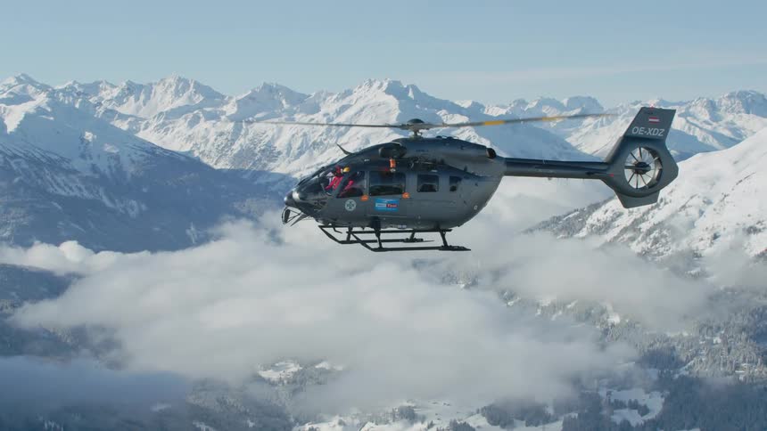 A militaristicappearing helicopter is shown from the side as it is flying above a wispy cloud over a snowcovered mountain range