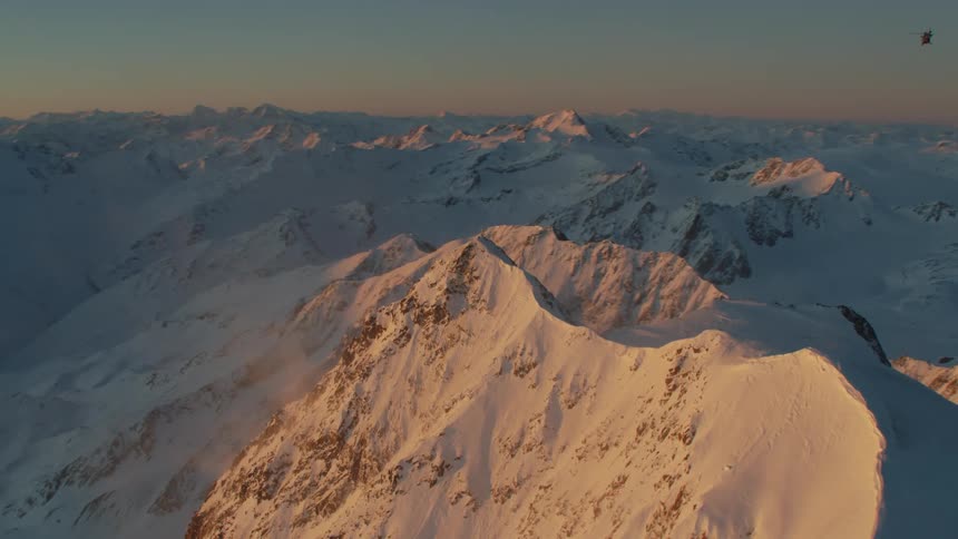 Helicopter flies around summit topped with spire in mountain range covered by snow and shadow
